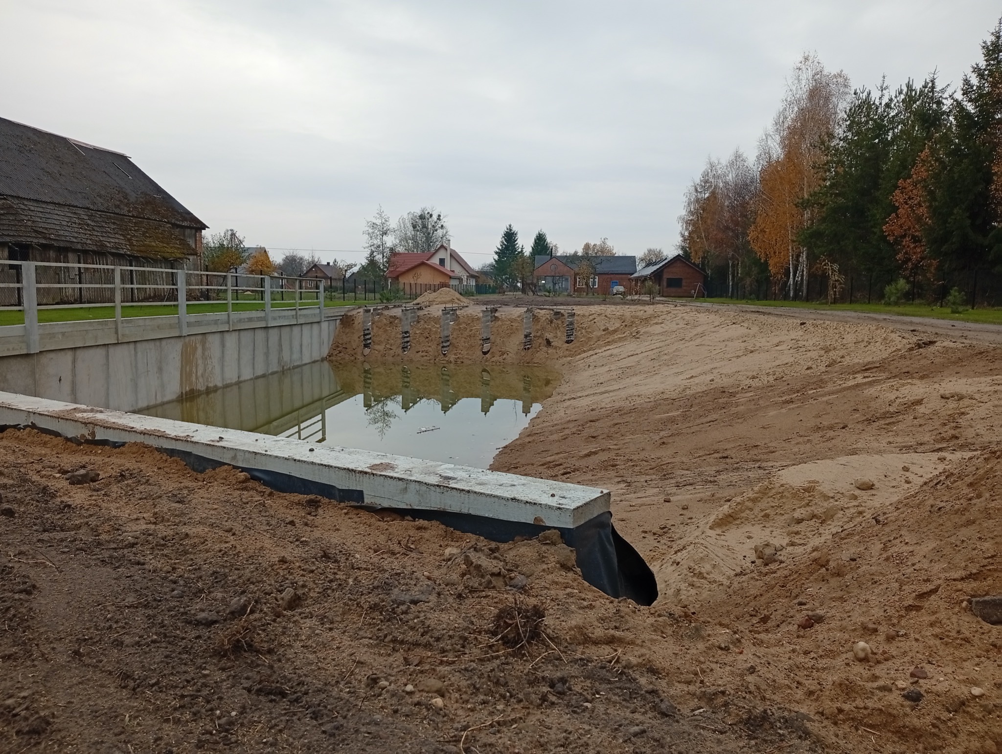 Budowa zbiornika retencyjnego w krajobrazie wiejskim. Widoczne betonowe elementy konstrukcyjne, ziemia i woda. W tle zabudowania i drzewa.
