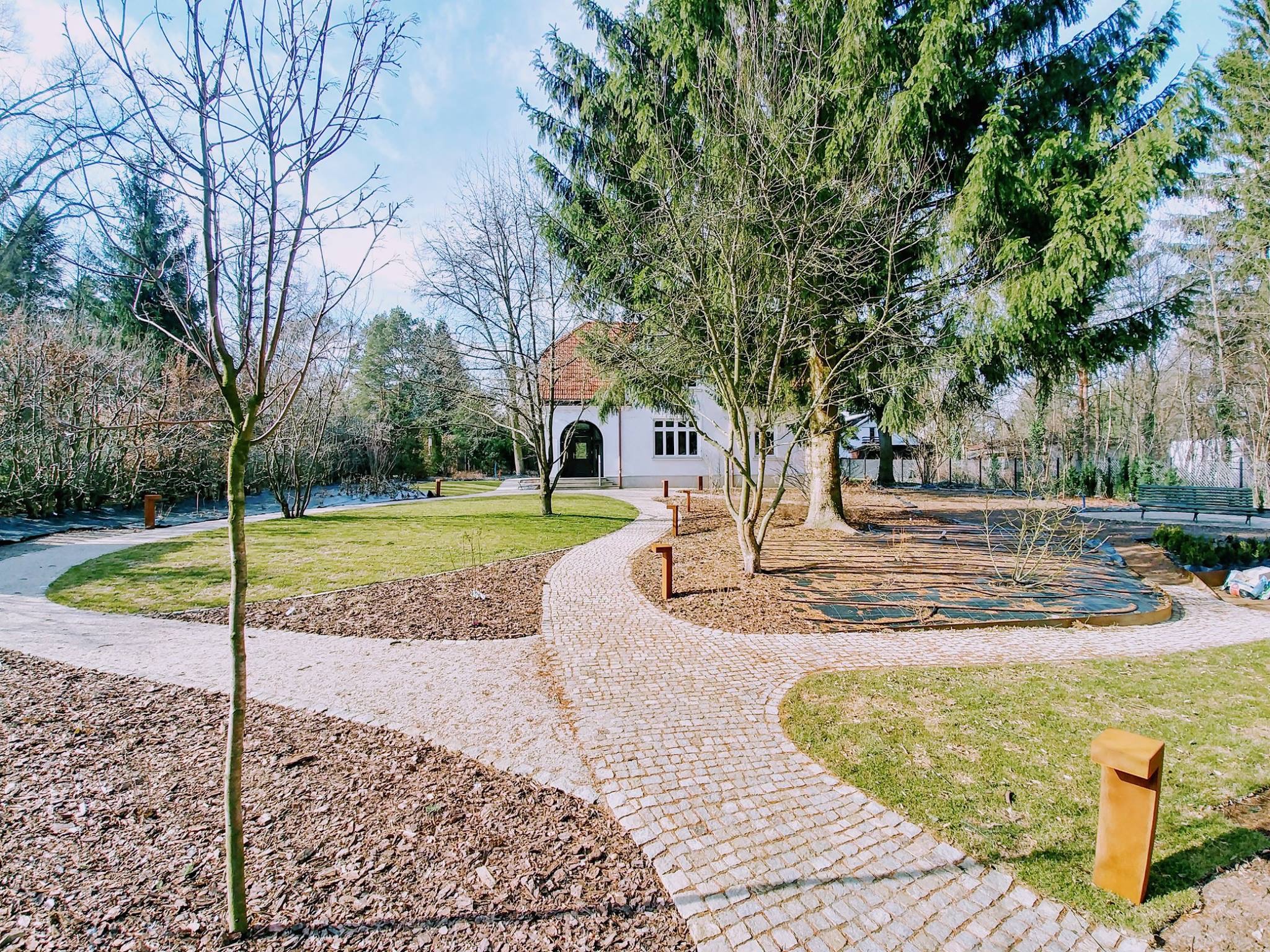 Biały budynek z czerwonym dachem widziany z perspektywy ścieżki z kostki brukowej, otoczony zielenią i drzewami. Elementy małej architektury.