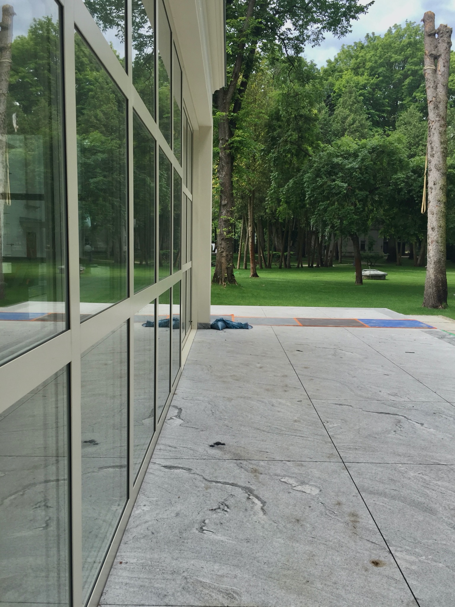 Nowoczesna elewacja budynku z dużymi oknami, odbijającymi zieleń ogrodu. Szara kamienna posadzka i minimalistyczny design. Widok z zewnątrz, w tle drzewa.