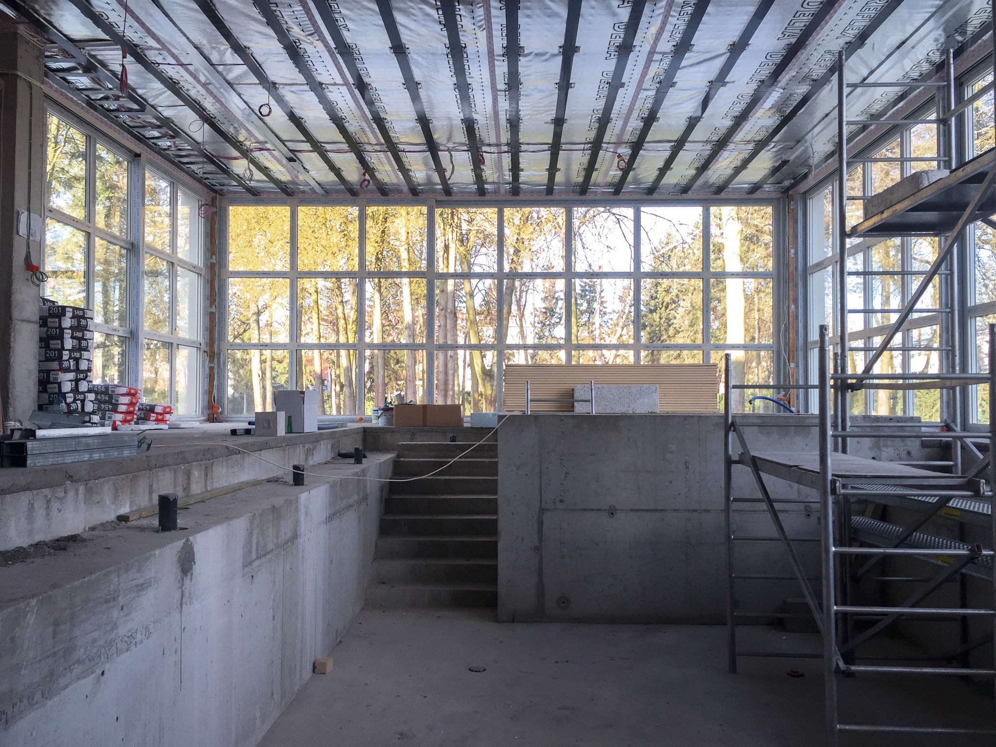 Wnętrze budynku w trakcie budowy z dużymi oknami, widoczne elementy konstrukcyjne sufitu, ściany z betonu, schody i rusztowanie. Prace wykończeniowe w surowym stanie.