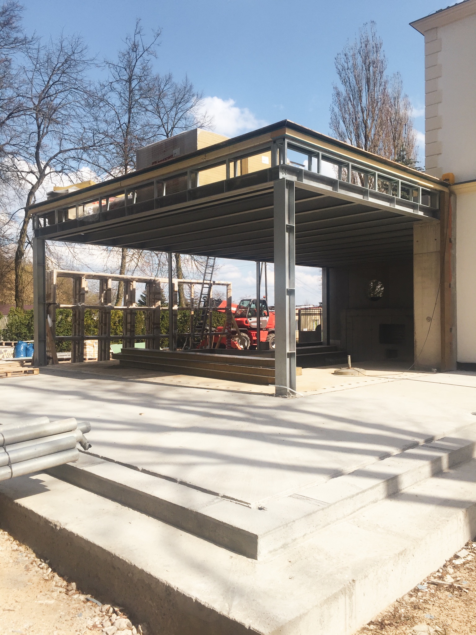 Konstrukcja stalowa zadaszenia w trakcie budowy, widoczny podnośnik i elementy drewniane. Jasne światło słoneczne, częściowo zacienione podłoże.