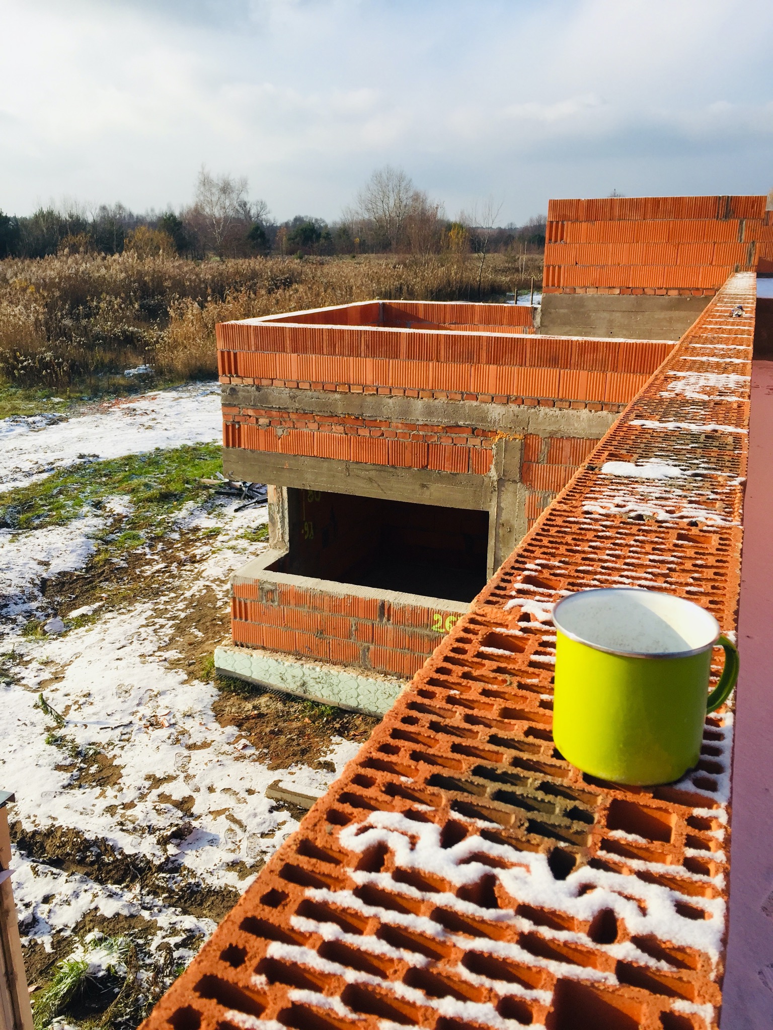 Wznoszenie murów z czerwonej cegły ceramicznej w surowym stanie, z widocznym fragmentem zielonego kubka na pierwszym planie i zimowym krajobrazem w tle.