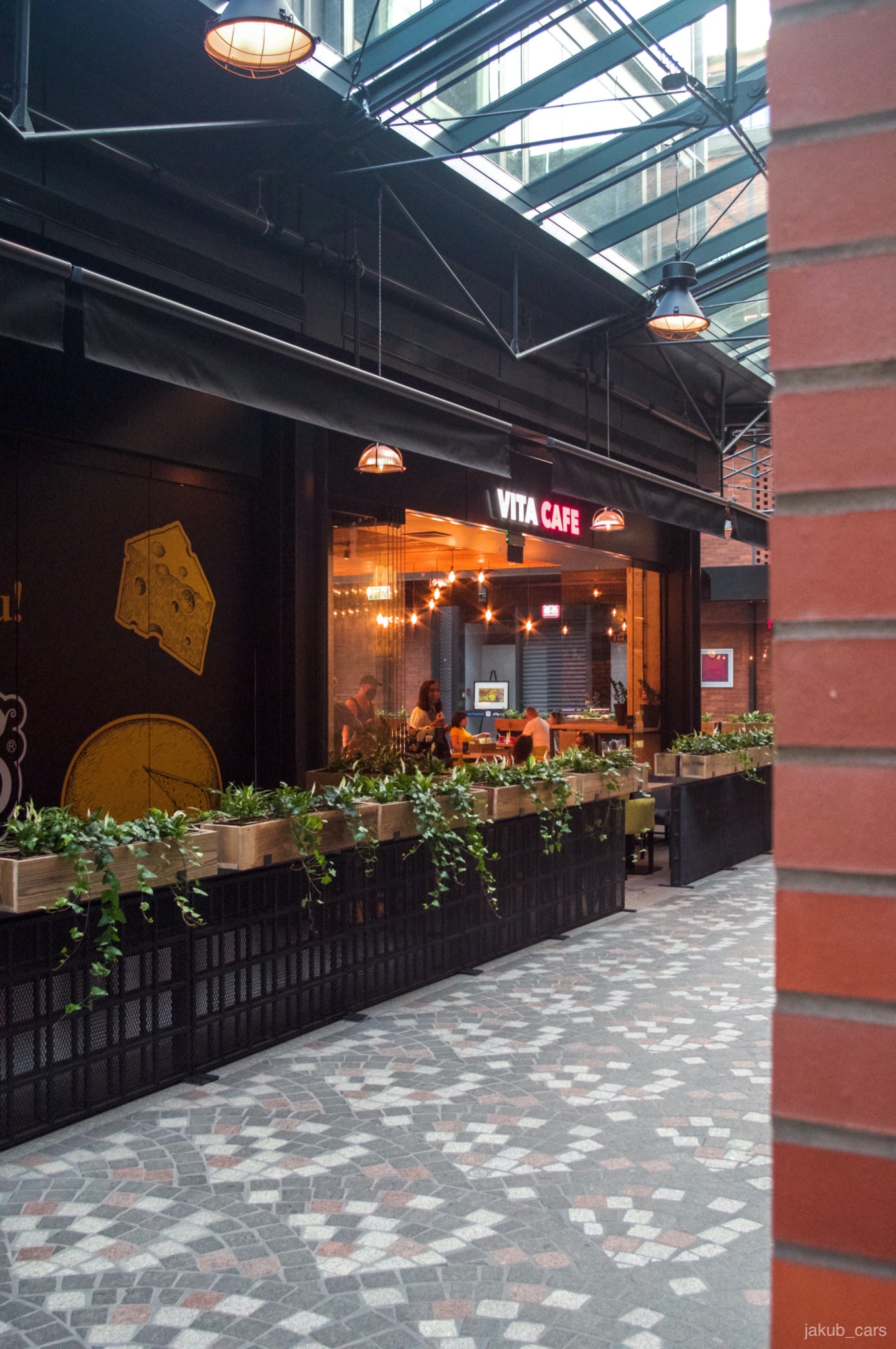 Elewacja budynku usługowego Vita Cafe z dekoracyjnymi elementami, roślinami w donicach i oświetleniem, widok z zewnątrz na chodnik z mozaiką.