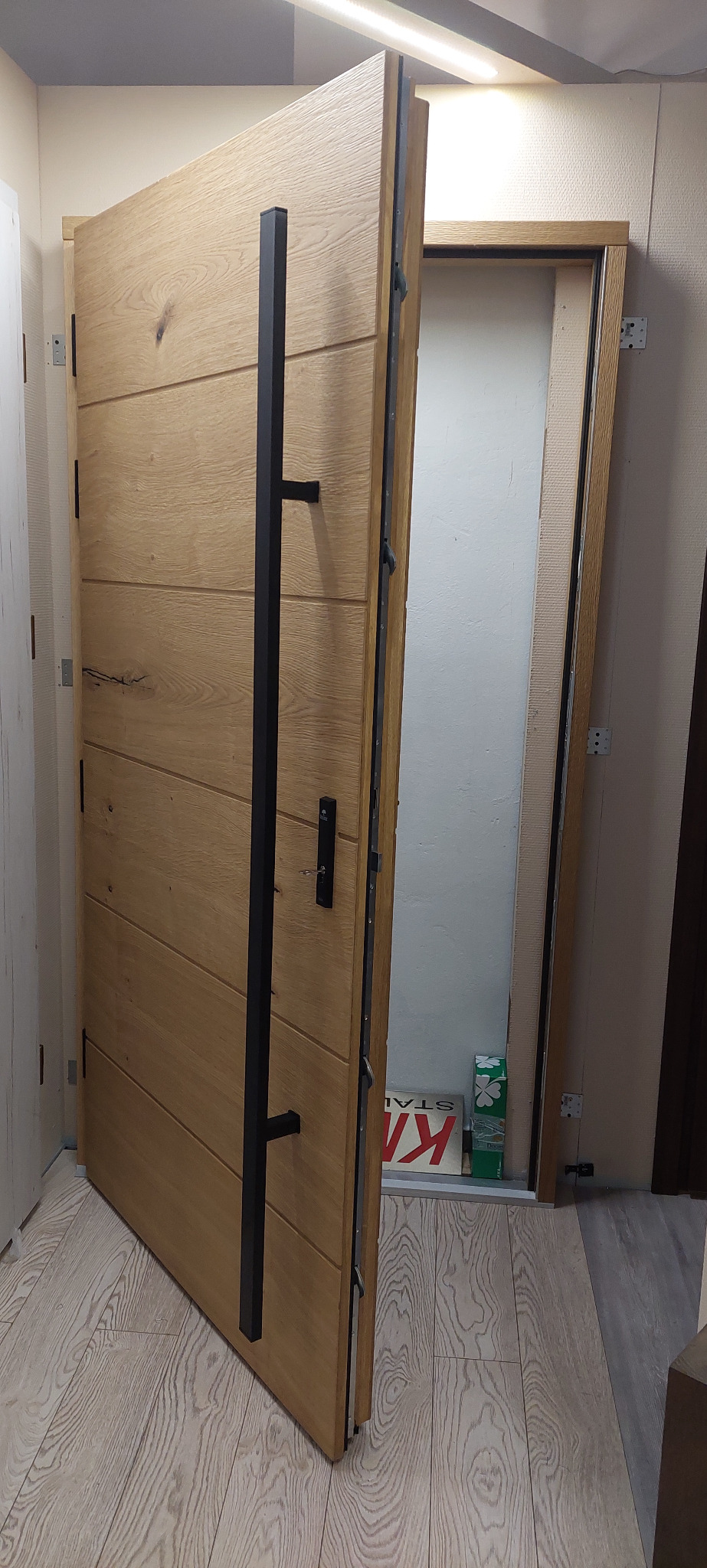 Otwarta dębowa drzwi z długim, czarnym, kwadratowym uchwytem, widoczna ościeżnica i podłoga w odcieniach bieli i szarości.