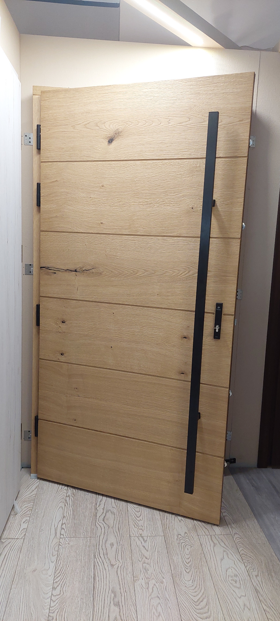 Nowoczesne, dębowe drzwi wejściowe z długim, czarnym uchwytem, zamontowane w jasnym wnętrzu, widoczne zawiasy i zamek.