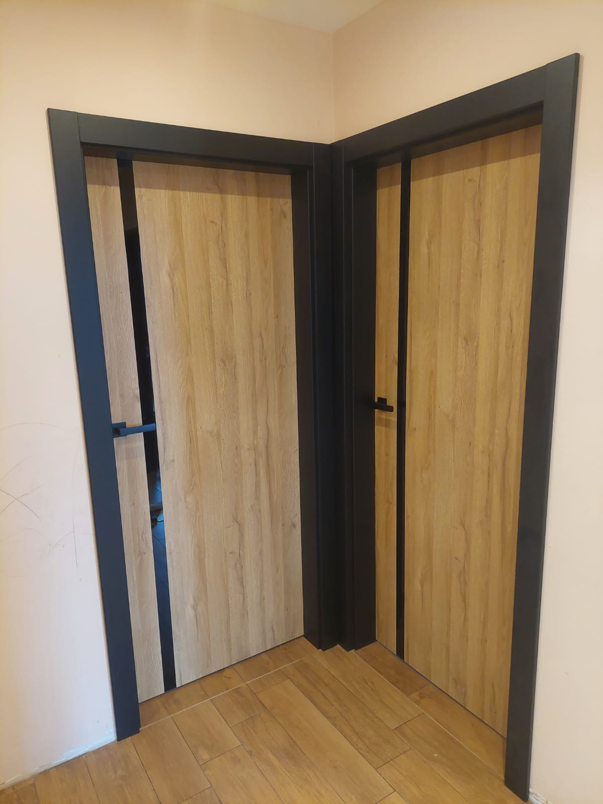 Dwa zamontowane, drewnopodobne drzwi wewnętrzne z czarnymi ościeżnicami w rogu pokoju, jeden uchylony, ukazujący fragment podłogi i ściany za nim.