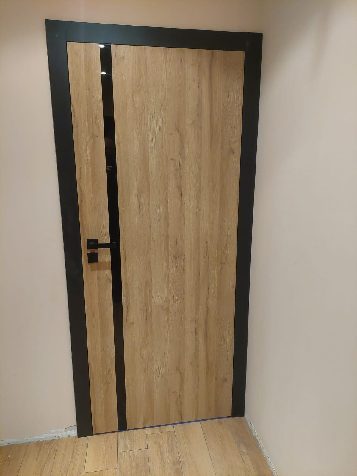 Zamknięte drzwi wewnętrzne z drewnopodobnym panelem, czarną ramą i pionowym paskiem po lewej stronie, klamka po lewej stronie, widoczne fragmenty ściany i podłogi.