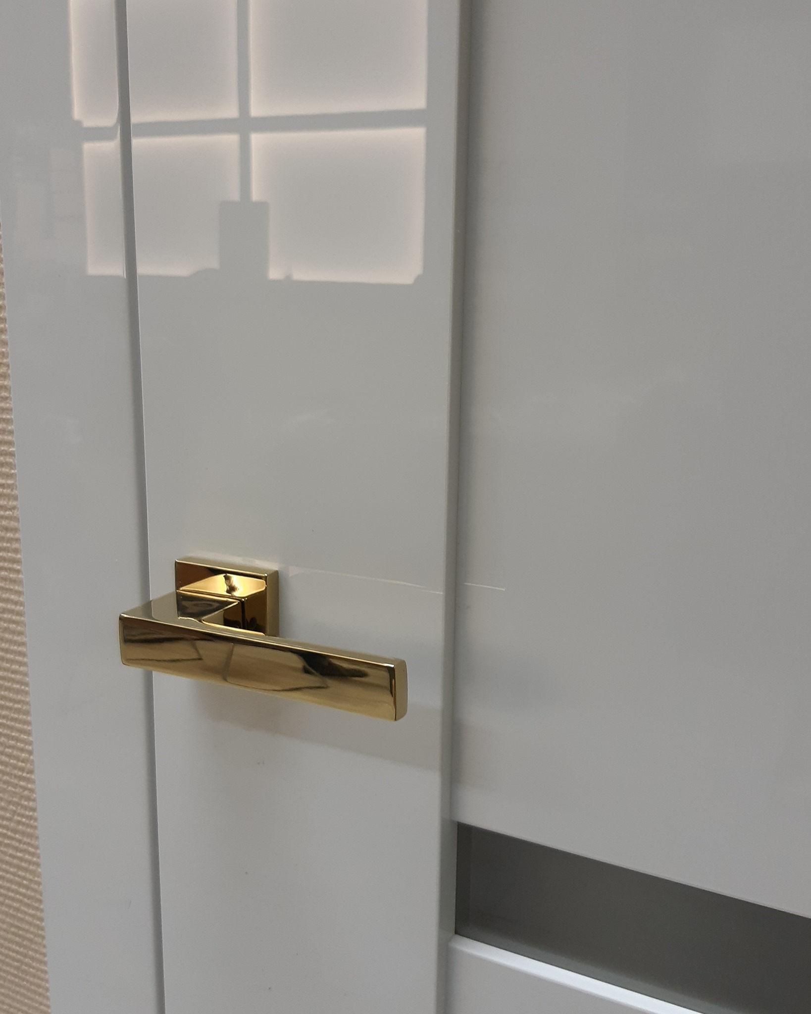 Białe, lakierowane drzwi wewnętrzne z poziomą, szarą wstawką i złotą klamką, odbijające światło okna.