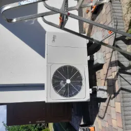 Zewnętrzna jednostka klimatyzacyjna Midea zamontowana na białej ścianie budynku, widoczna drabina oparta o ścianę, kostka brukowa na ziemi.