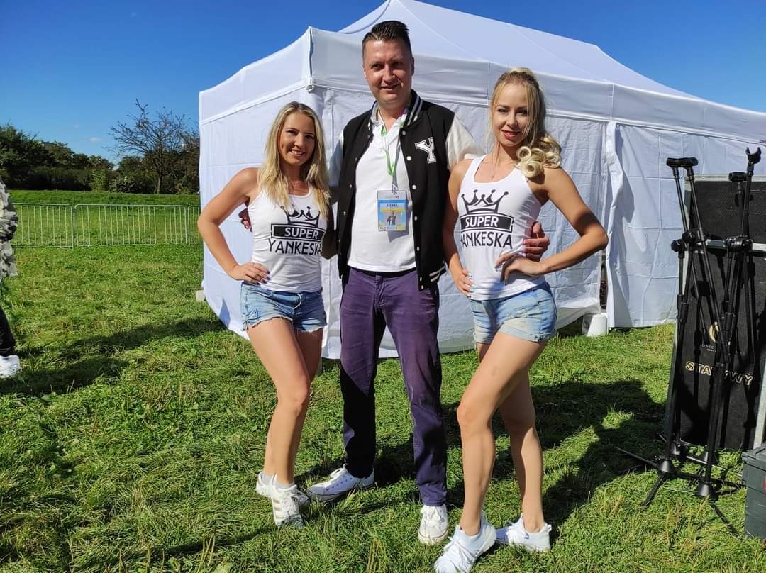 Trzy osoby pozują na trawie przed białym namiotem: dwie kobiety w białych topach z napisem 'SUPER YANKESKA' i dżinsowych szortach oraz mężczyzna w kurtce bejsbolówce i spodniach w kolorze śliwkowym...