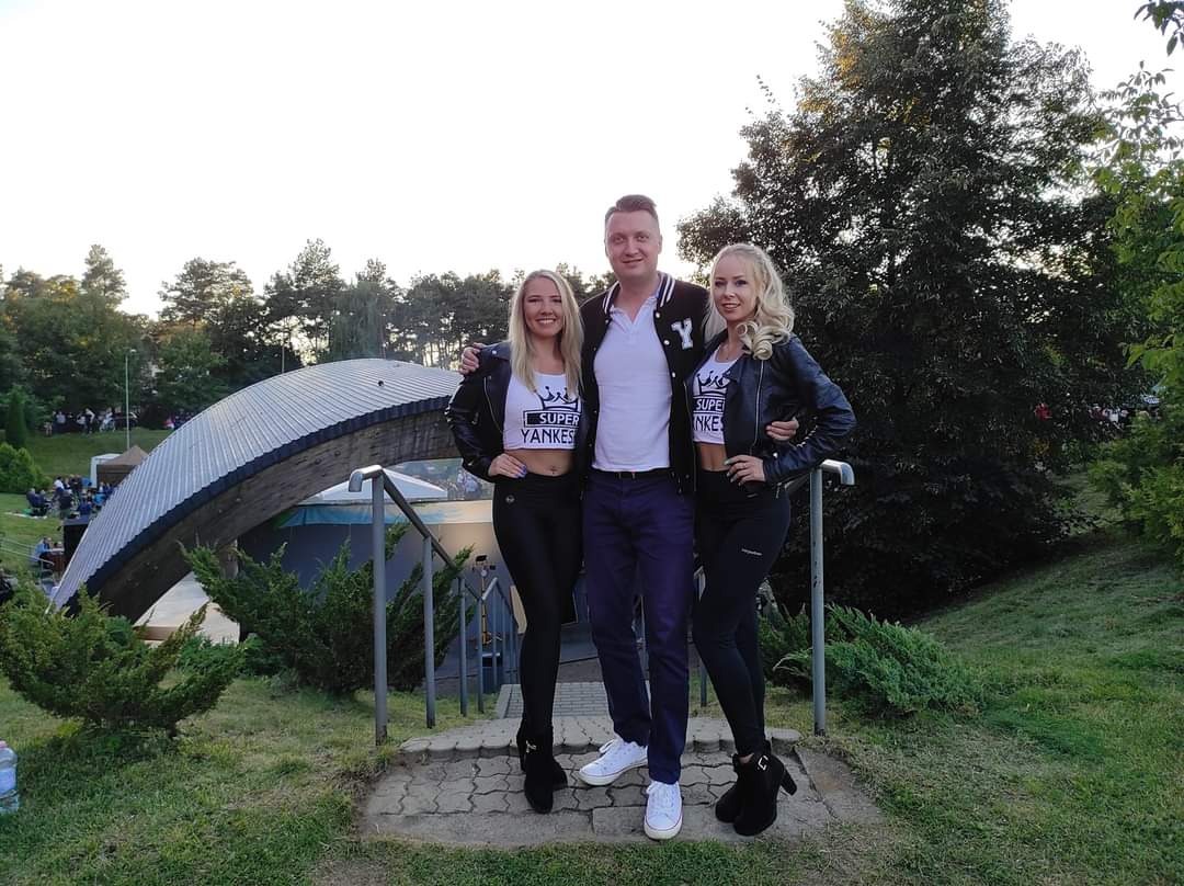 Trzy osoby pozują na schodach na tle plenerowej sceny z zadaszeniem; dwie kobiety w czarnych legginsach i skórzanych kurtkach, w białych topach z nadrukiem 'Super Yankees', oraz mężczyzna...