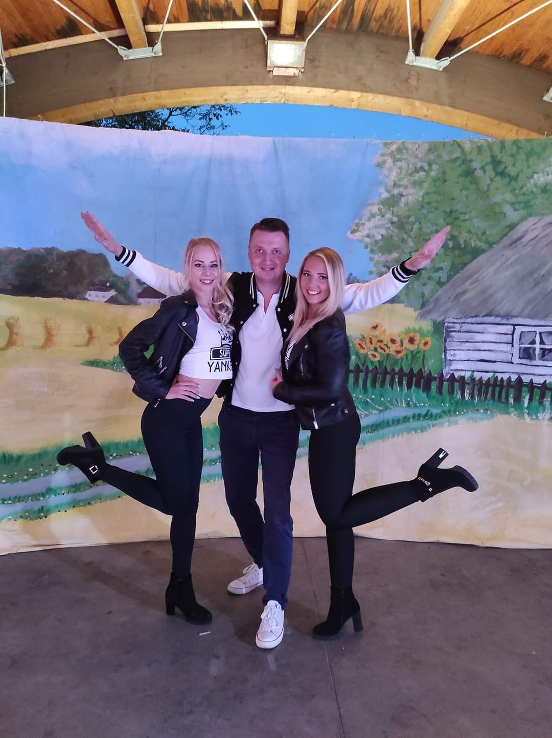 Zespół muzyczny pozuje przed ręcznie malowaną scenografią przedstawiającą wiejski krajobraz z chatą i słonecznikami.