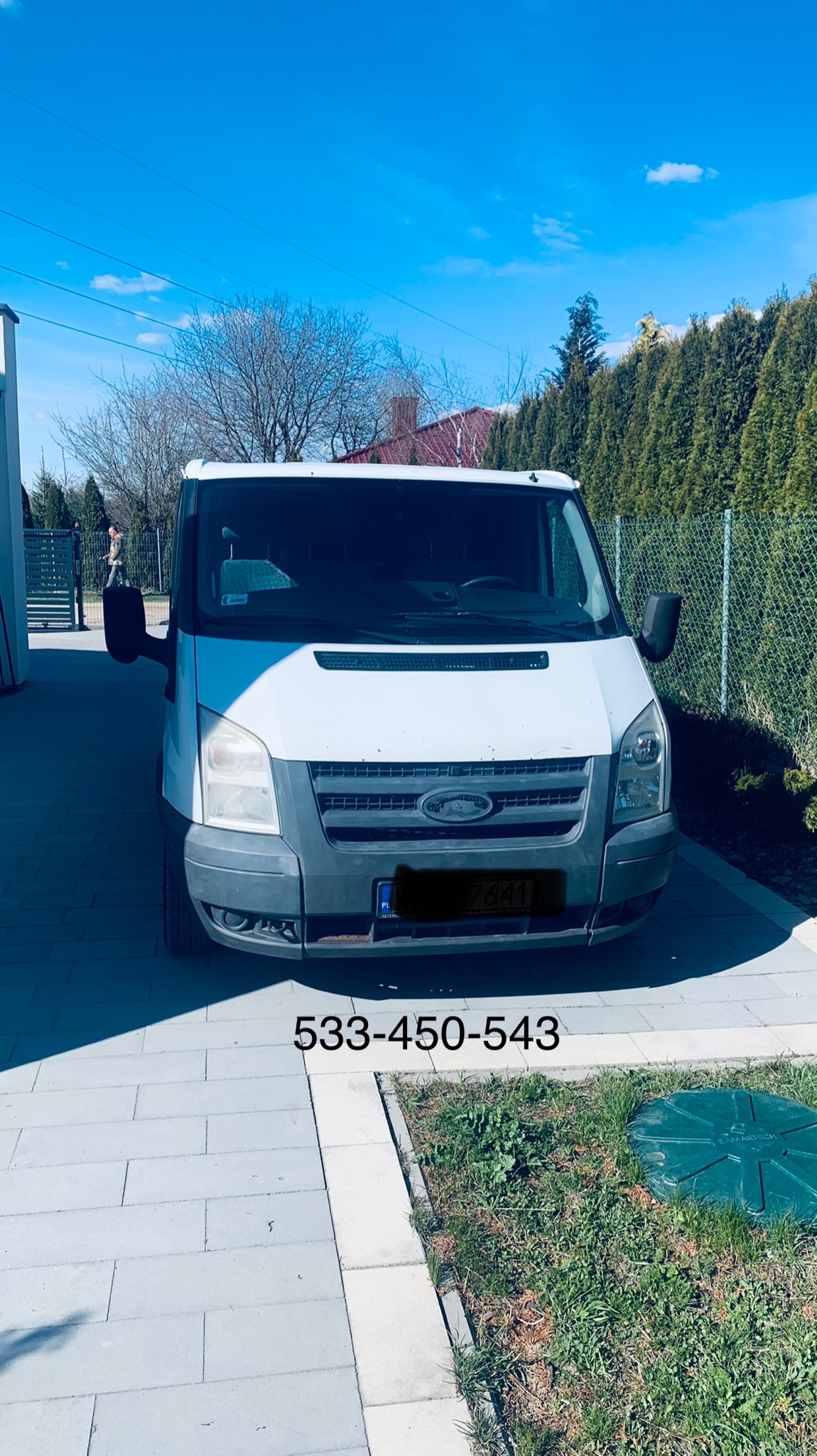 Biały Ford Transit zaparkowany na podjeździe z kostki brukowej w słoneczny dzień, widoczny numer telefonu kontaktowego na zdjęciu, w tle zielony żywopłot.