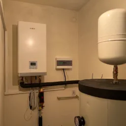 Biały wiszący piec gazowy marki Aiwa z elektronicznym wyświetlaczem, podłączony do instalacji, obok biały zbiornik z czarnym blatem i manometrem, na tle jasnych ścian.