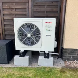 Biała jednostka zewnętrzna pompy ciepła Aiwa Inverter R32, zamontowana na betonowych bloczkach z czarnymi podkładkami, ustawiona przed brązowym drewnianym płotem i beżową ścianą budynku.