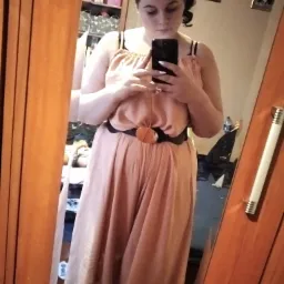 Kobieta robi selfie w lustrze, ubrana w długi, luźny kombinezon w odcieniu brzoskwini z czarnym paskiem i okrągłą klamrą. W tle widoczne zdjęcia w ramkach na ścianie i regał z różnymi przedmiotami.