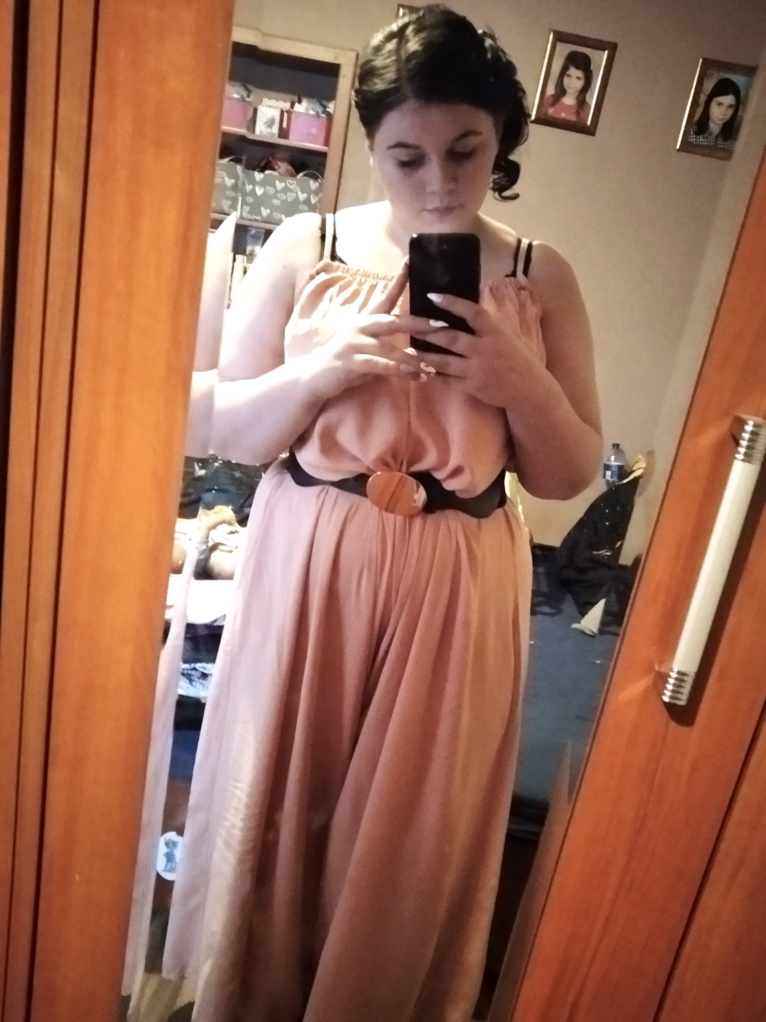 Kobieta robi selfie w lustrze, ubrana w długi, luźny kombinezon w odcieniu brzoskwini z czarnym paskiem i okrągłą klamrą. W tle widoczne zdjęcia w ramkach na ścianie i regał z różnymi przedmiotami.