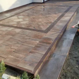 Ułożona kostka brukowa w odcieniach brązu tworzy wzór na tarasie z dwoma stopniami. Widoczne rynny i fragment zieleni. Ciekawy układ kostki w centralnej części tarasu.