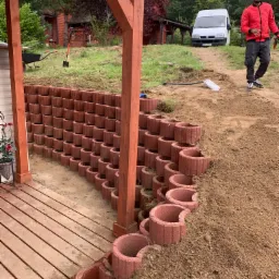 Budowa murku oporowego z czerwonych, łukowatych elementów betonowych, widoczny pracownik w czerwonej kurtce, teren w trakcie prac ziemnych, w tle dom i biały dostawczy samochód.