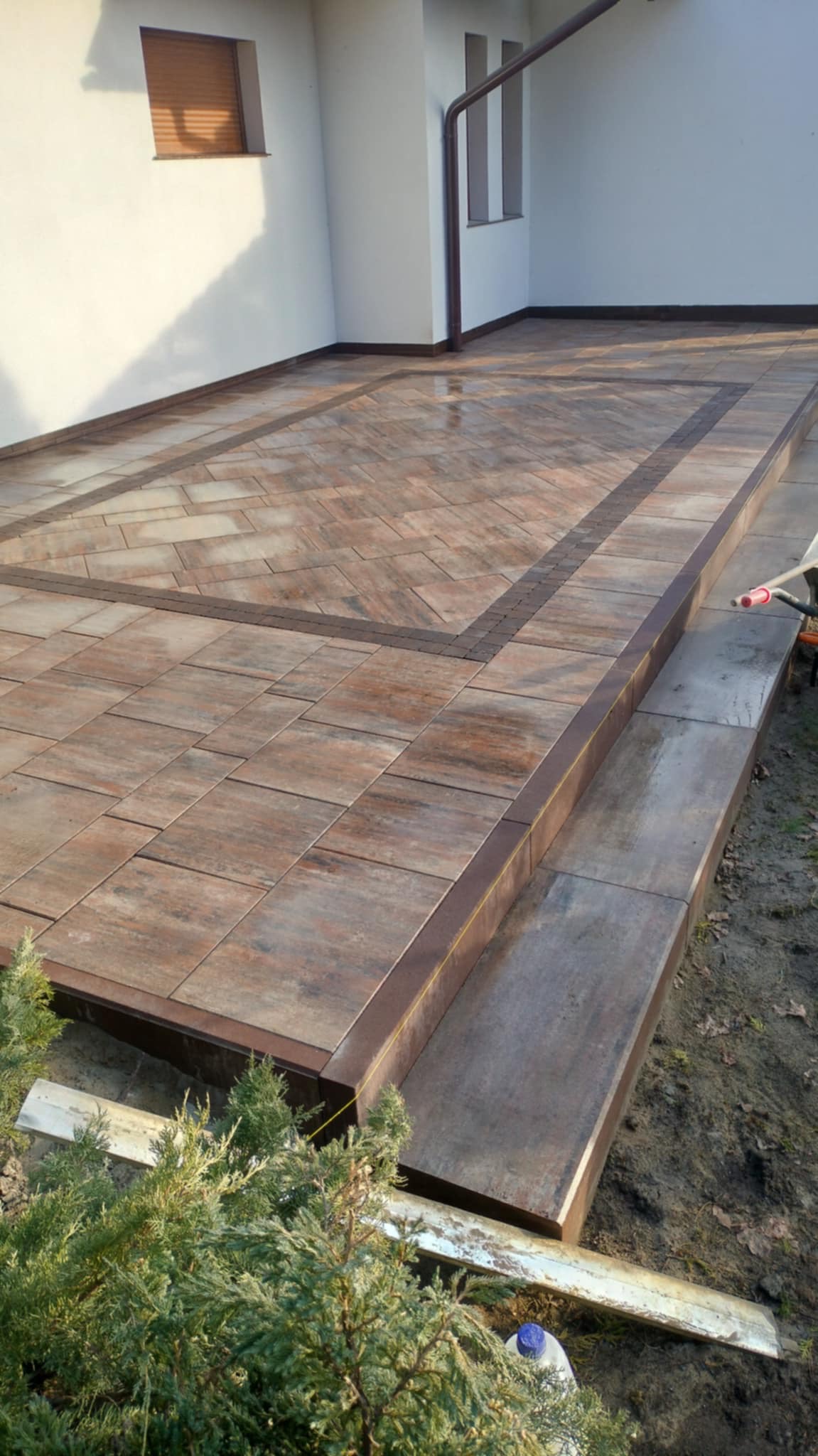 Ułożona kostka brukowa w odcieniach brązu tworzy wzór na tarasie z dwoma stopniami. Widoczne rynny i fragment zieleni. Ciekawy układ kostki w centralnej części tarasu.