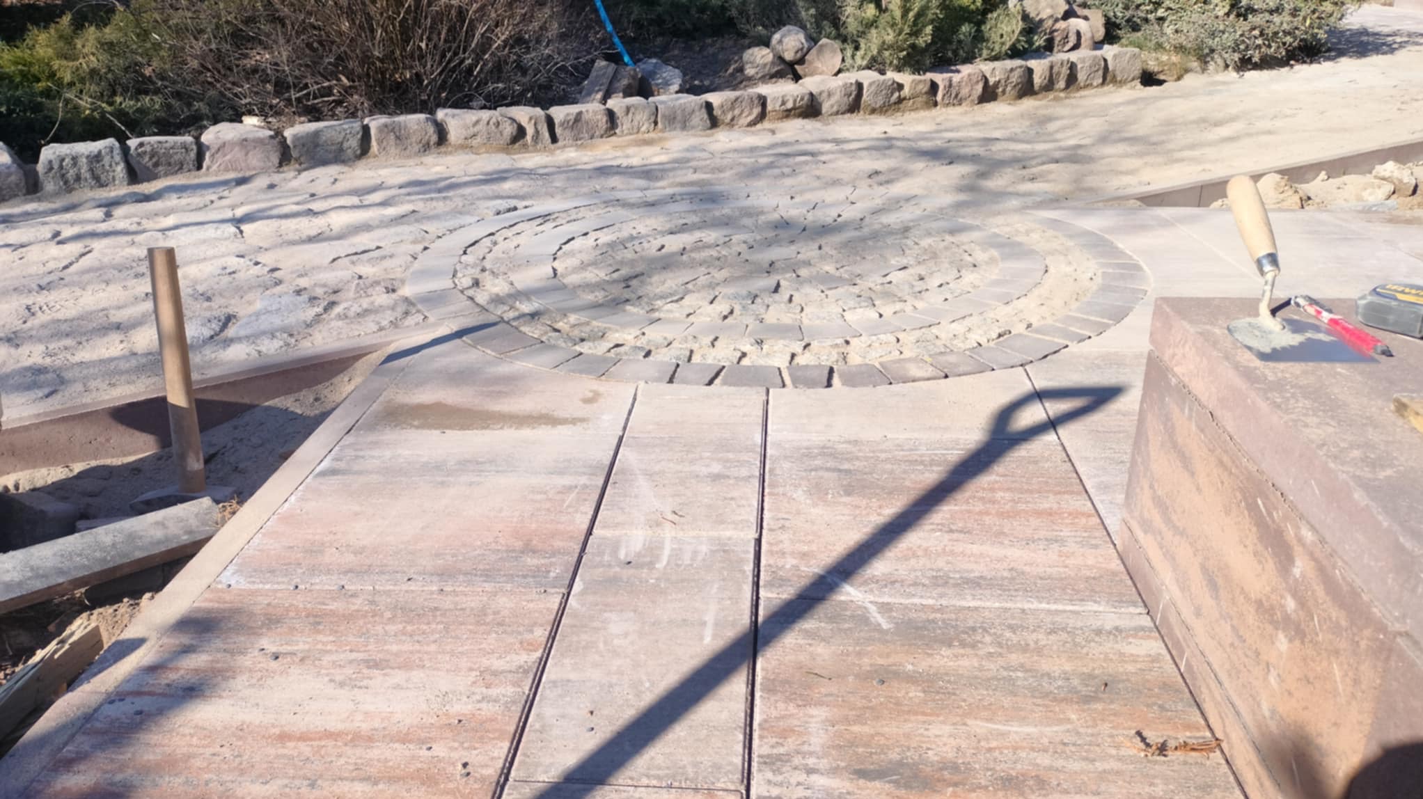 Kremowo-brązowa kostka brukowa w trakcie układania, widoczny okrągły wzór i narzędzia brukarskie, w tle roślinność i kamienne obrzeże.