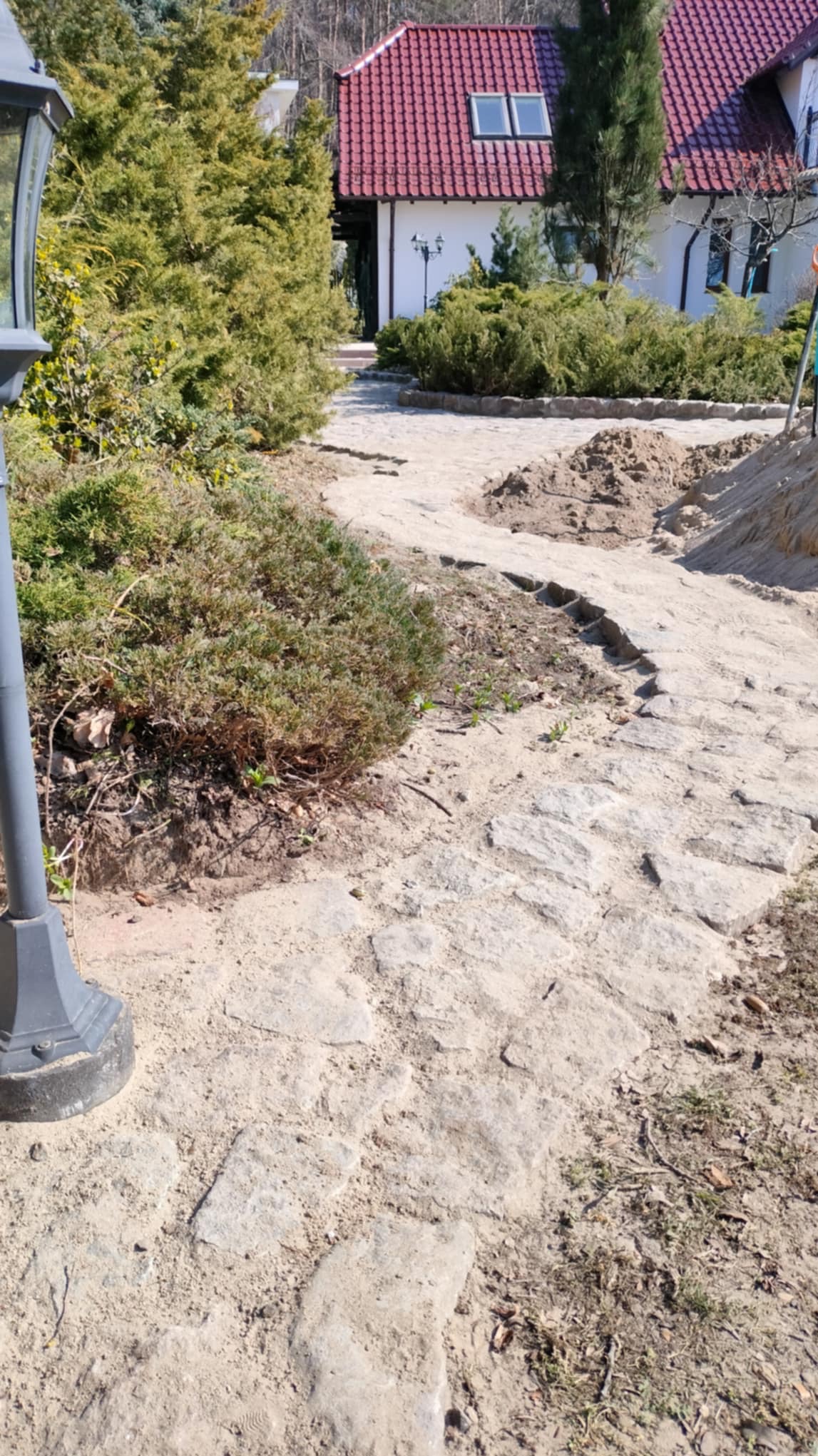 Ścieżka z kamieni w ogrodzie, częściowo zasypana piaskiem, z widocznymi krzewami i domem w tle. Lampa ogrodowa po lewej stronie kadru. Prace w toku.