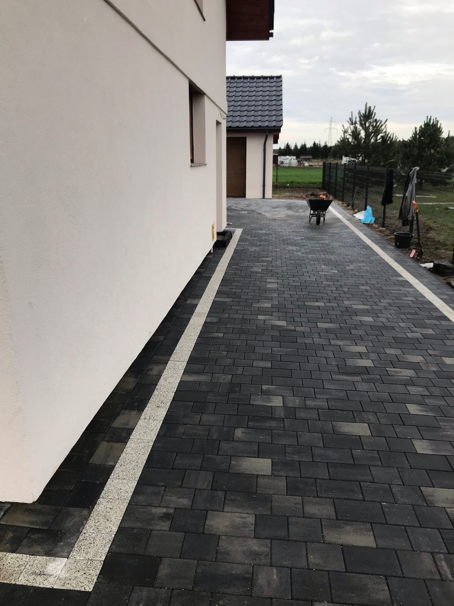 Ułożona ciemnoszara kostka brukowa z białym pasem oddzielającym ją od ściany budynku, taczka z ziemią w tle.