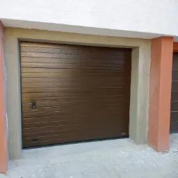 Zamknięta, pozioma, brązowa brama garażowa segmentowa z małym zamkiem po lewej stronie, zamontowana w surowej, nieotynkowanej wnęce, z widoczną kostką brukową na podjeździe.