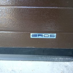 Stolarka otworowa 4K - Brązowy panel bramy garażowej z wytłoczonym wzorem i niebieskim logo 'GROS', widoczne uszczelnienie dolne i fragment betonowej posadzki.