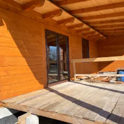 Drewniany dom w trakcie budowy: widoczne okna z brązowymi ramami, drewniana elewacja pomalowana na pomarańczowo i fragment drewnianego tarasu w surowym stanie.