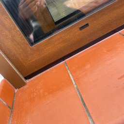 Stolarka otworowa 4K - Fragment okna PCV w kolorze drewna, detale uszczelnienia i połączenia z terakotą. Widoczne odbicie osoby robiącej zdjęcie.