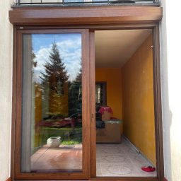 Stolarka otworowa 4K - Brązowe, przesuwne drzwi balkonowe z roletą zewnętrzną, widok na ogród z odbiciem w szybie. Pomarańczowe ściany wewnątrz balkonu.