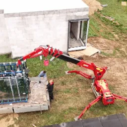 Montaż okien z użyciem dźwigu na budowie. Okna na samochodzie dostawczym, pracownicy nadzorują proces. Widok z góry na budynek w trakcie budowy.