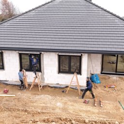 Stolarka otworowa 4K - Montaż okien z czarną ramą w surowym budynku. Trzech monterów przy pracy, drabiny, narzędzia. Widok z góry, na pierwszym planie ziemia.