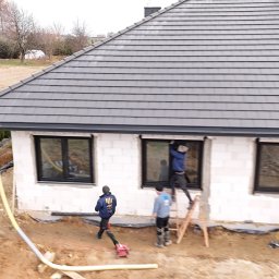 Stolarka otworowa 4K - Montaż okien w surowym budynku z białego bloczka. Ekipa na drabinie instaluje okno. Widoczne czarne ramy okienne i szare dachówki.