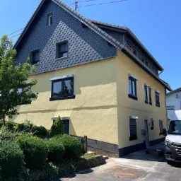 Żółty dom z szarym dachem krytym łuską, z zainstalowanymi roletami zewnętrznymi w kolorze szarym, widok z zewnątrz w słoneczny dzień.