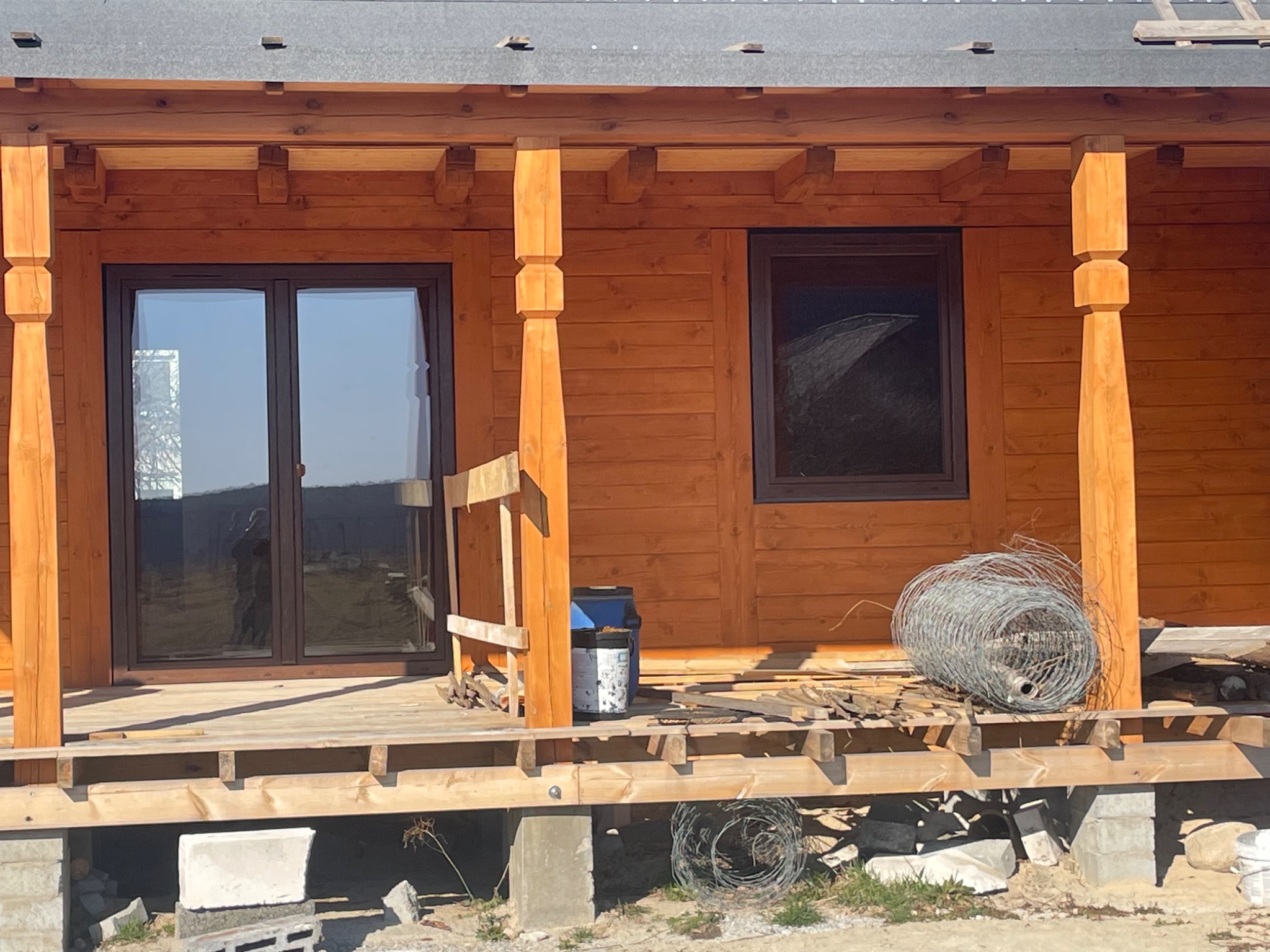 Drewniany dom z brązowymi oknami, w trakcie budowy werandy. Widoczne materiały budowlane i narzędzia. Częściowo wykończona konstrukcja.