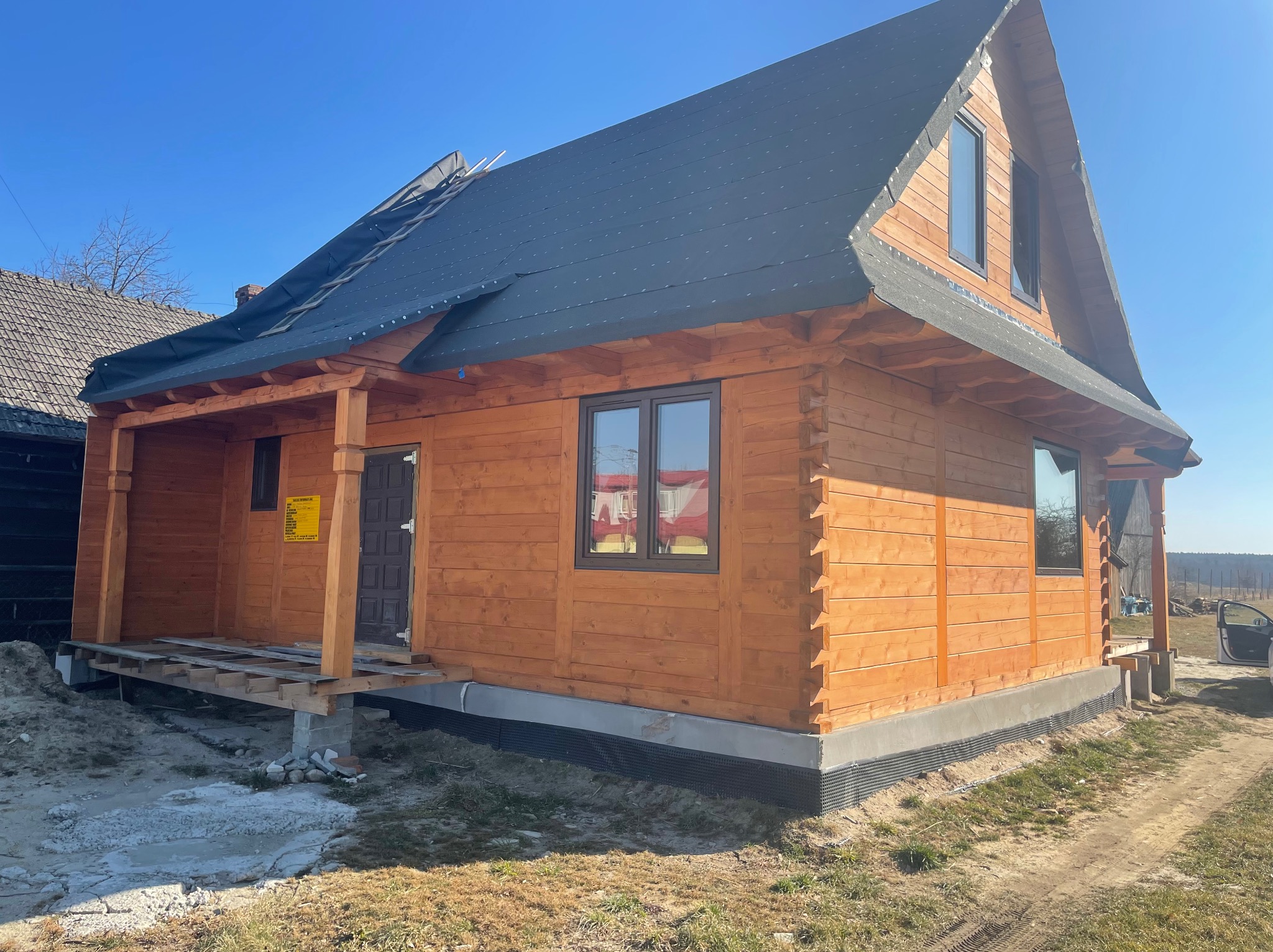 Surowa drewniana konstrukcja domu z oknami i dachem pokrytym papą, w słoneczny dzień. Widoczny fragment niezabudowanego tarasu i drabina oparta o dach.