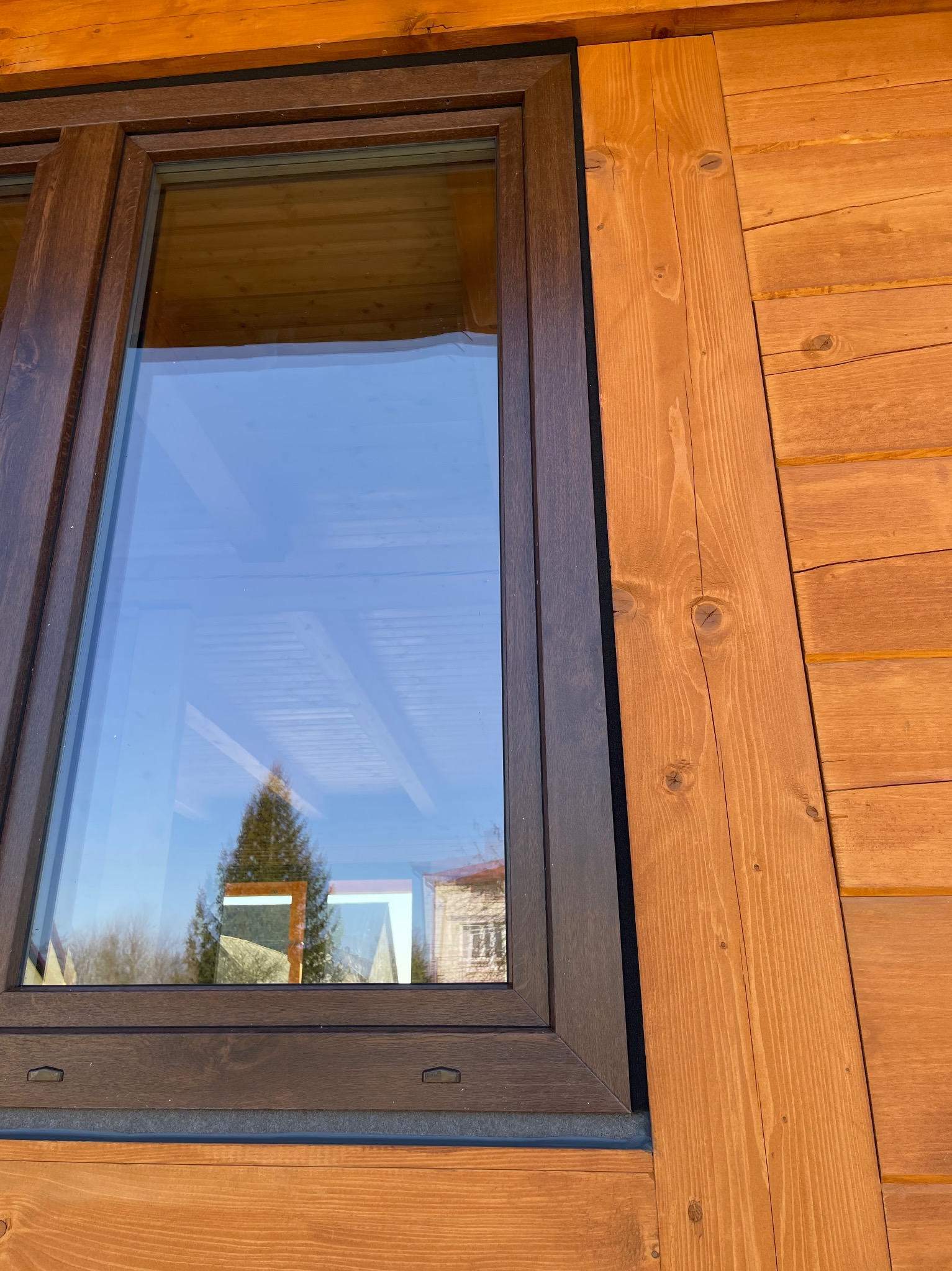 Okno z brązową ramą w drewnianej elewacji. Widoczne detale wykończenia i refleksy otoczenia w szybie. Charakterystyczny węzeł w desce elewacyjnej.