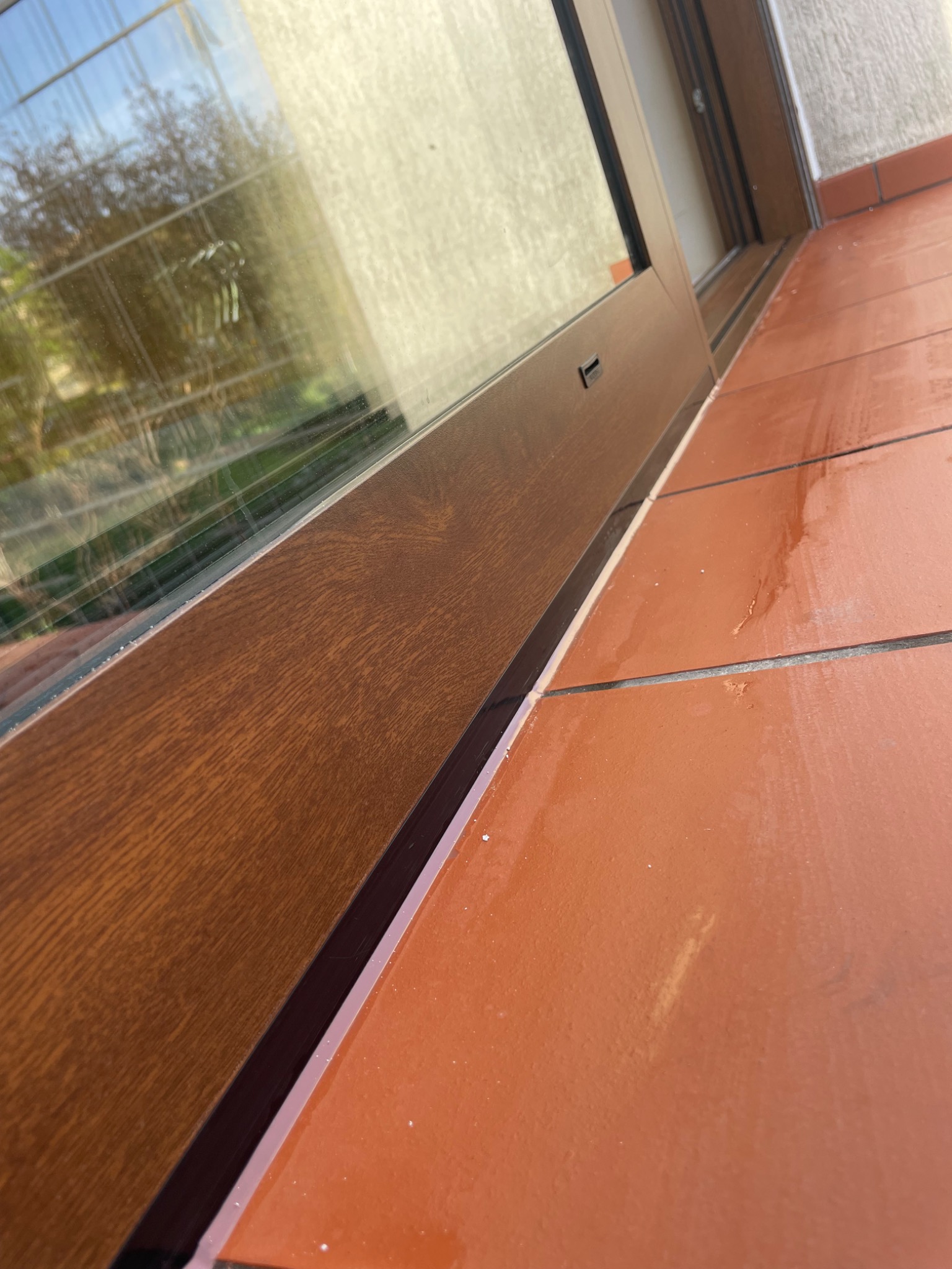 Brązowe okno z widocznymi detalami ramy i szybą odbijającą otoczenie, ułożone na pomarańczowych, błyszczących płytkach tarasowych.