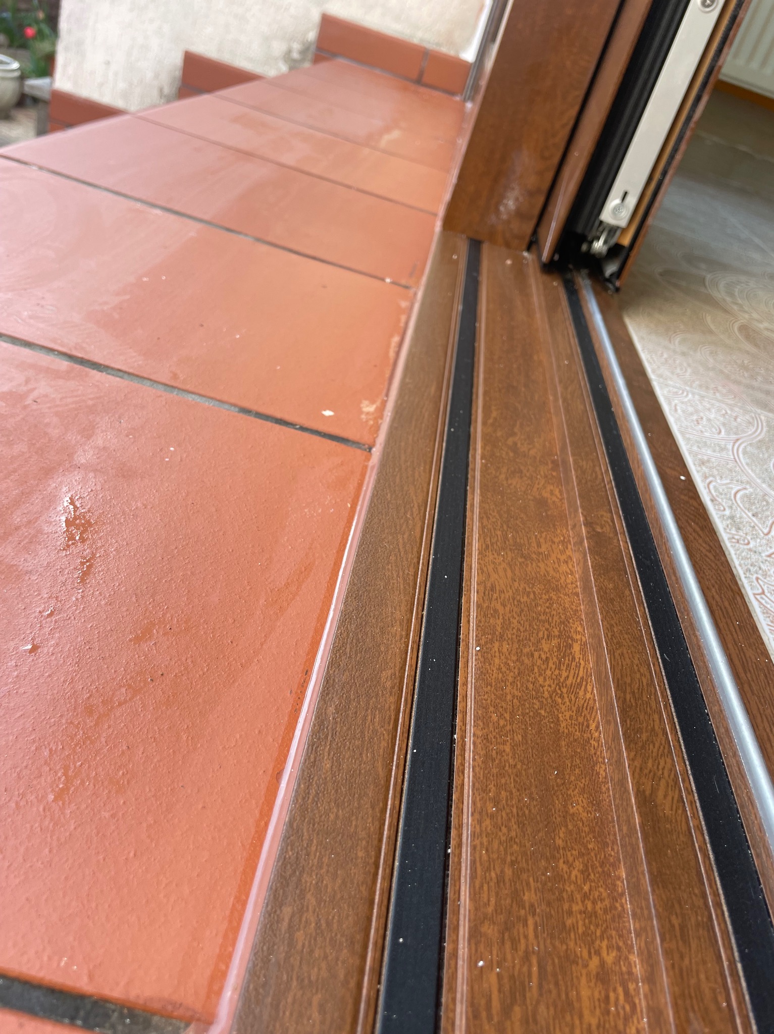 Niski kąt na brązowe drzwi przesuwne z widoczną uszczelką i fragmentem ceramicznych schodów wejściowych. Detal montażu, ciepłe odcienie drewna.