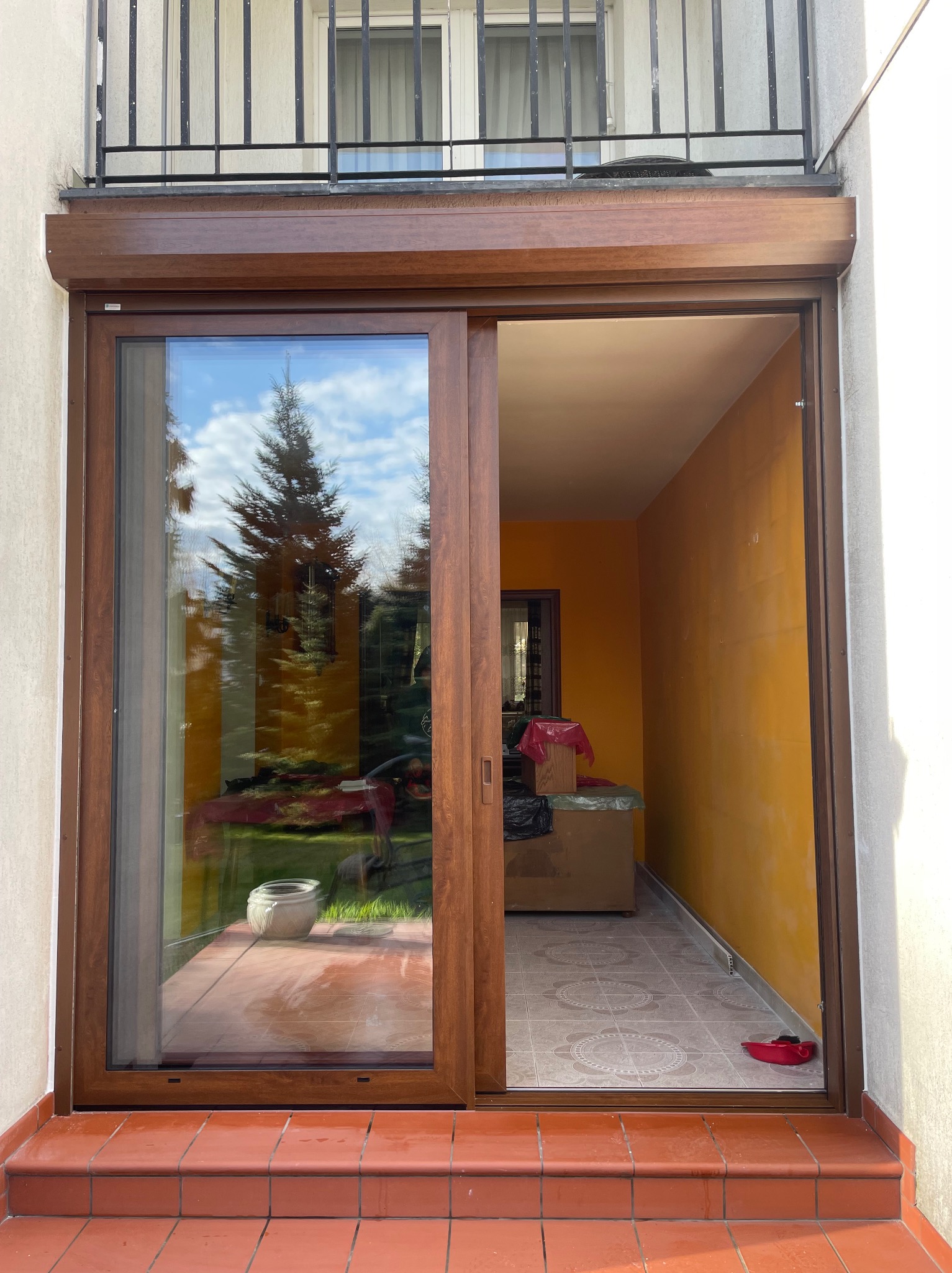Brązowe, przesuwne drzwi balkonowe z roletą zewnętrzną, widok na ogród z odbiciem w szybie. Pomarańczowe ściany wewnątrz balkonu.