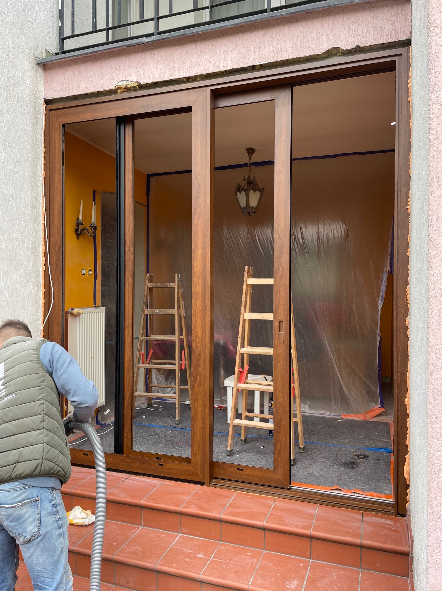 Montaż brązowych drzwi przesuwnych balkonowych. Wnętrze zabezpieczone folią malarską. Pracownik z odkurzaczem sprząta po montażu.