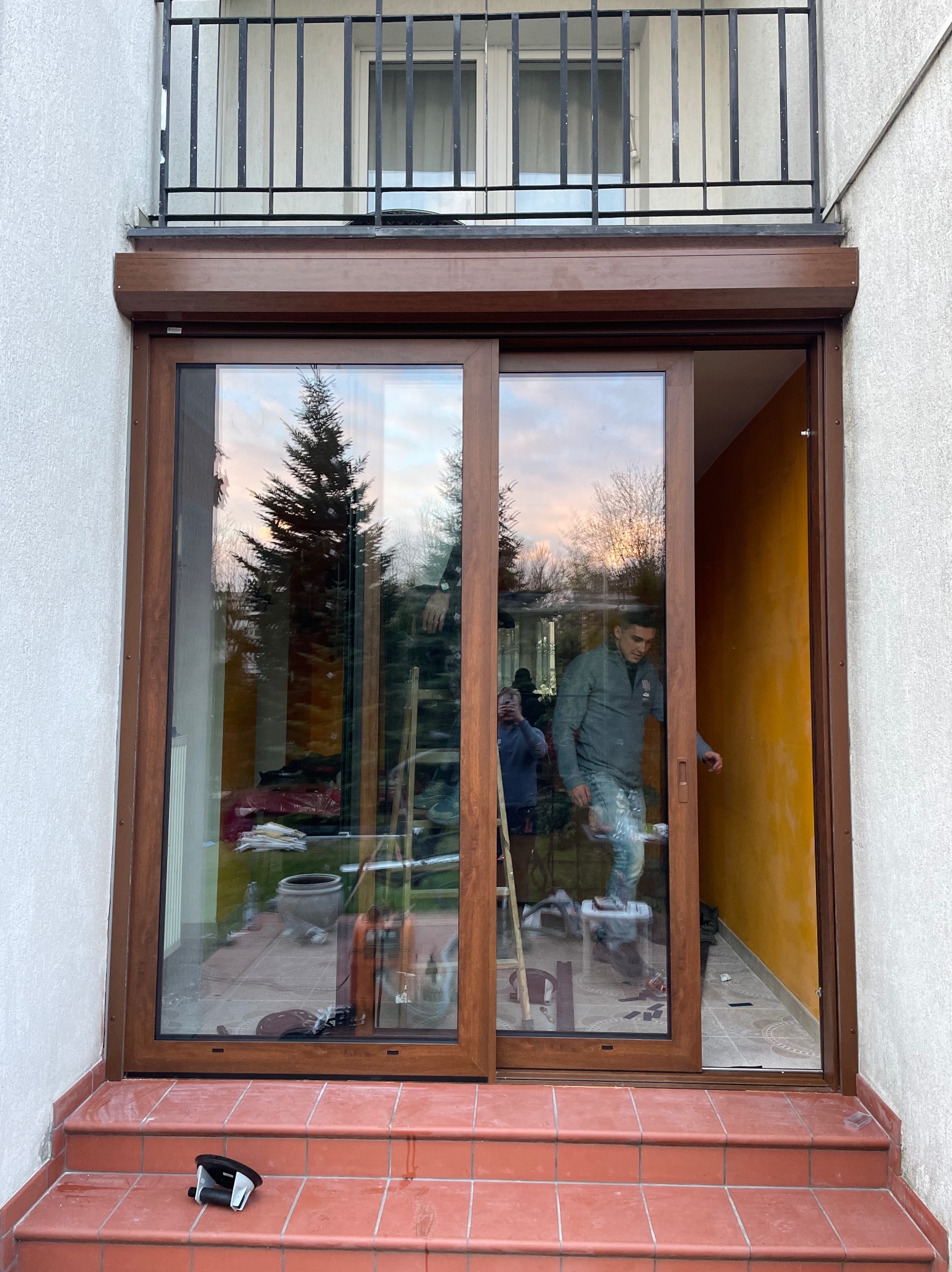 Montaż brązowych drzwi przesuwnych z roletą zewnętrzną w budynku z balkonem. Widoczni pracownicy podczas instalacji, narzędzia pracy. Pomarańczowa ściana w tle.