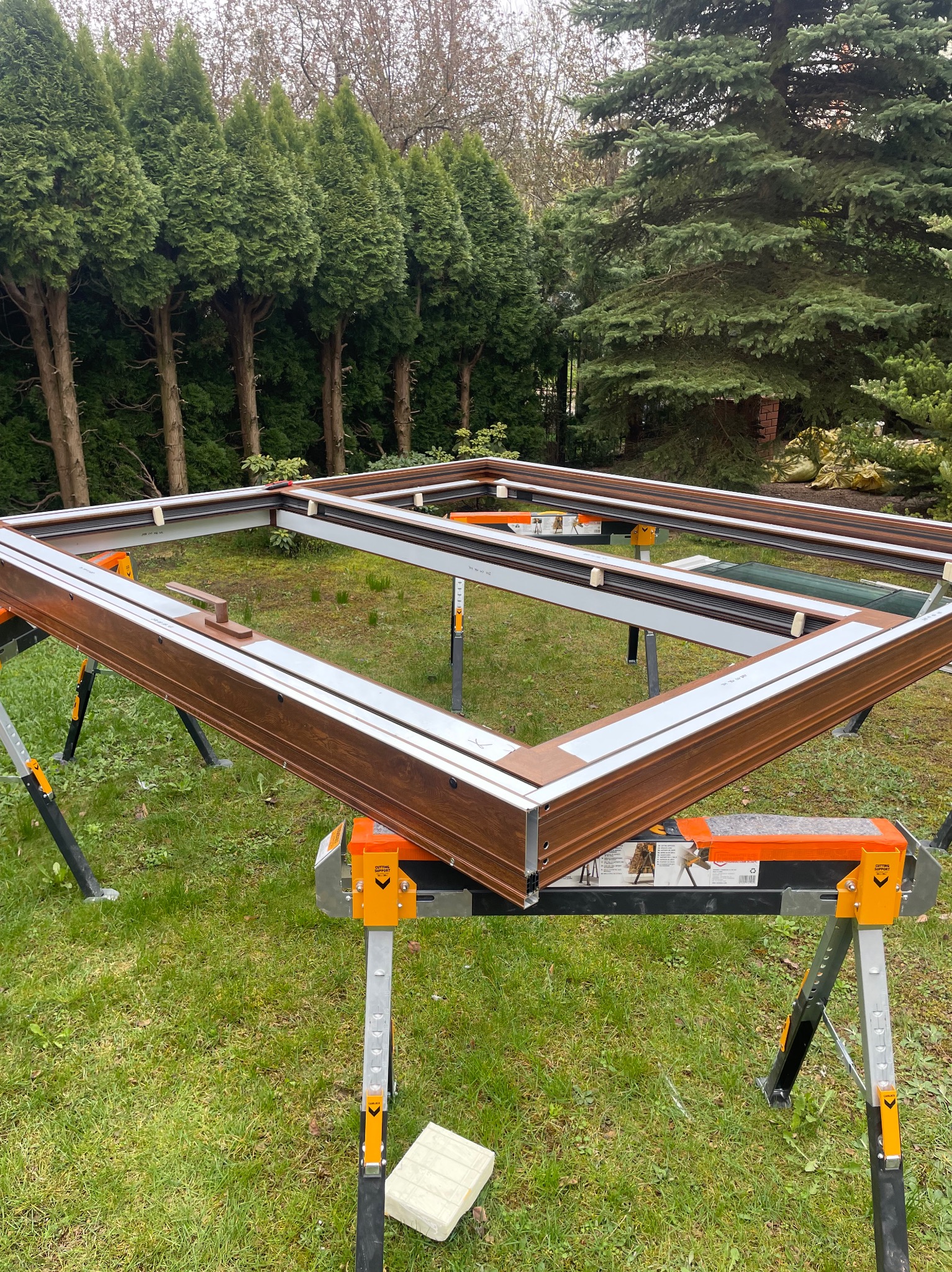 Brązowa rama okienna z szybą na kozłach montażowych na trawie, w tle zielone drzewa. Widoczne detale konstrukcyjne.