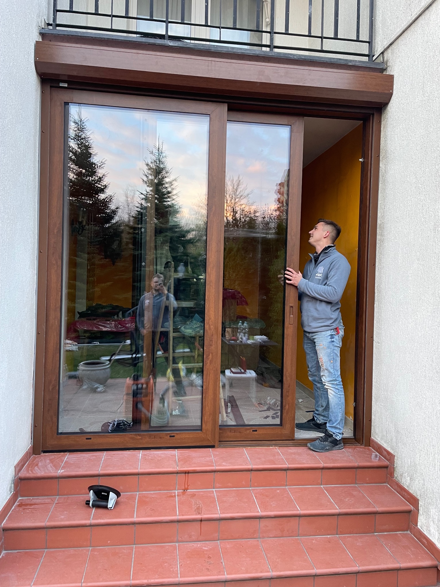 Montaż okna przesuwnego z brązową ramą. Mężczyzna w bluzie i jeansach kontroluje montaż. Na schodach leży przyssawka do szyb. Widoczna roleta.