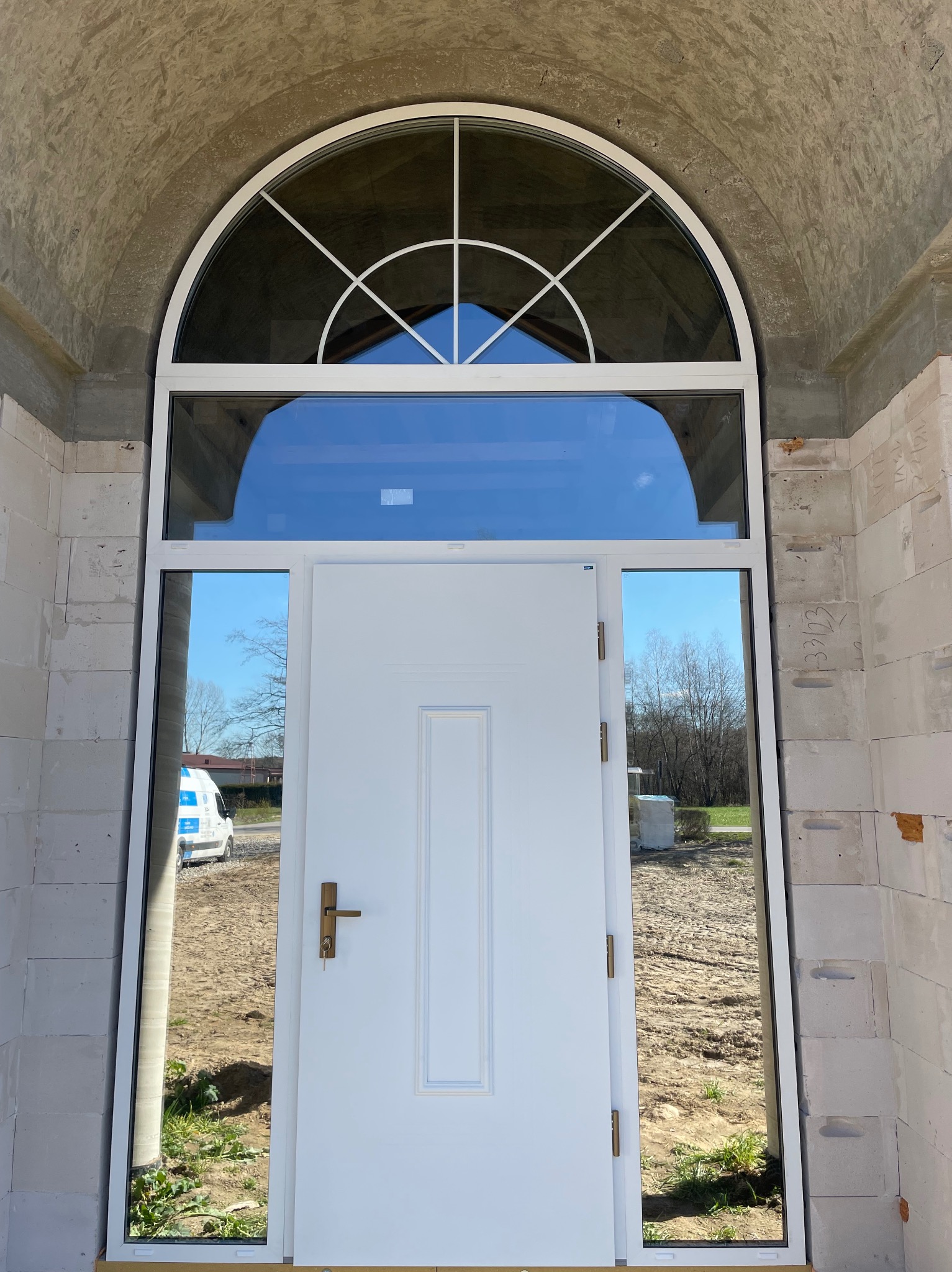 Białe drzwi wejściowe z półokrągłym oknem nadświetlnym, zamontowane w surowym murze. W szybach odbija się krajobraz i zaparkowany samochód.