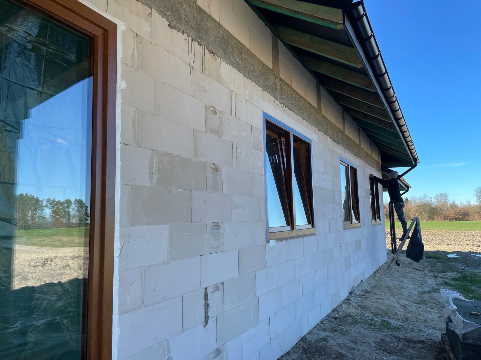 Montaż okien z brązowymi ramami w surowym budynku z betonu komórkowego. Widoczna osoba na drabinie, montująca rynnę. Błękitne niebo odbija się w szybach.