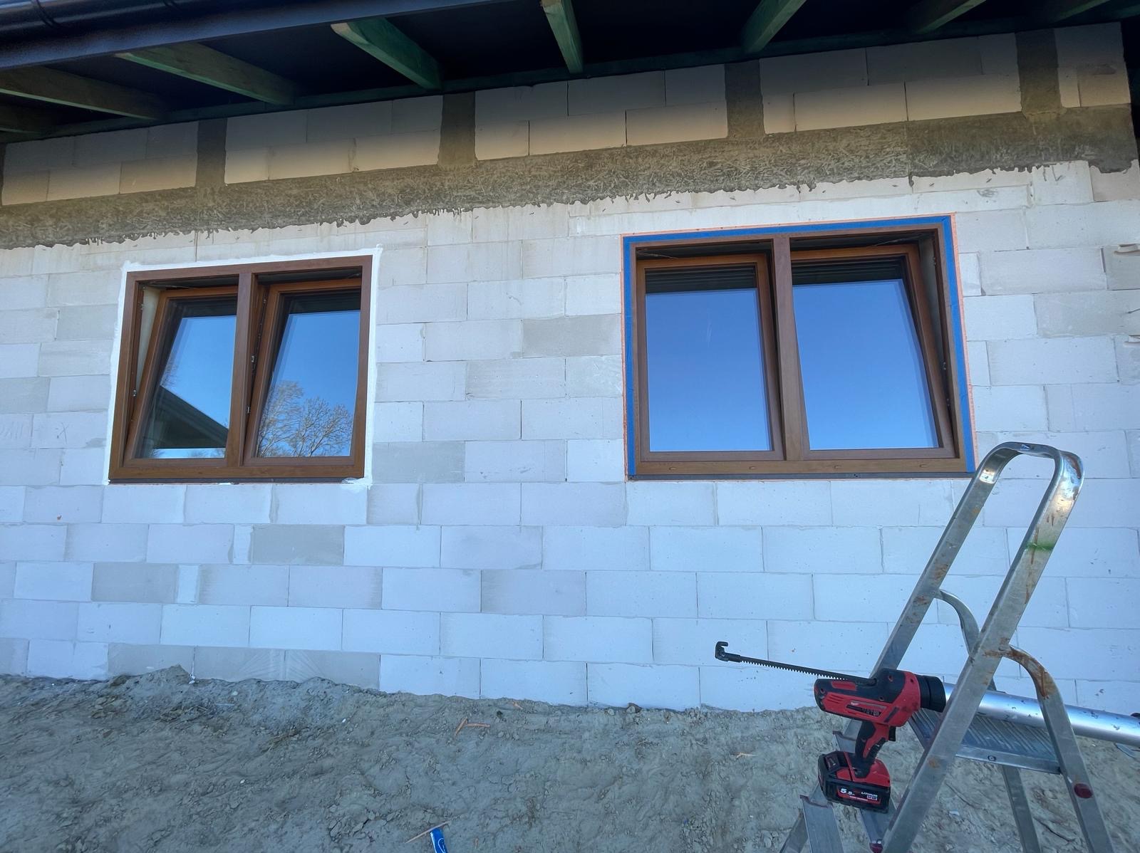 Montaż okien z brązowymi ramami w ścianie z białych bloczków. Widoczna drabina i narzędzie do uszczelniania. Jedno okno z niebieską taśmą zabezpieczającą.