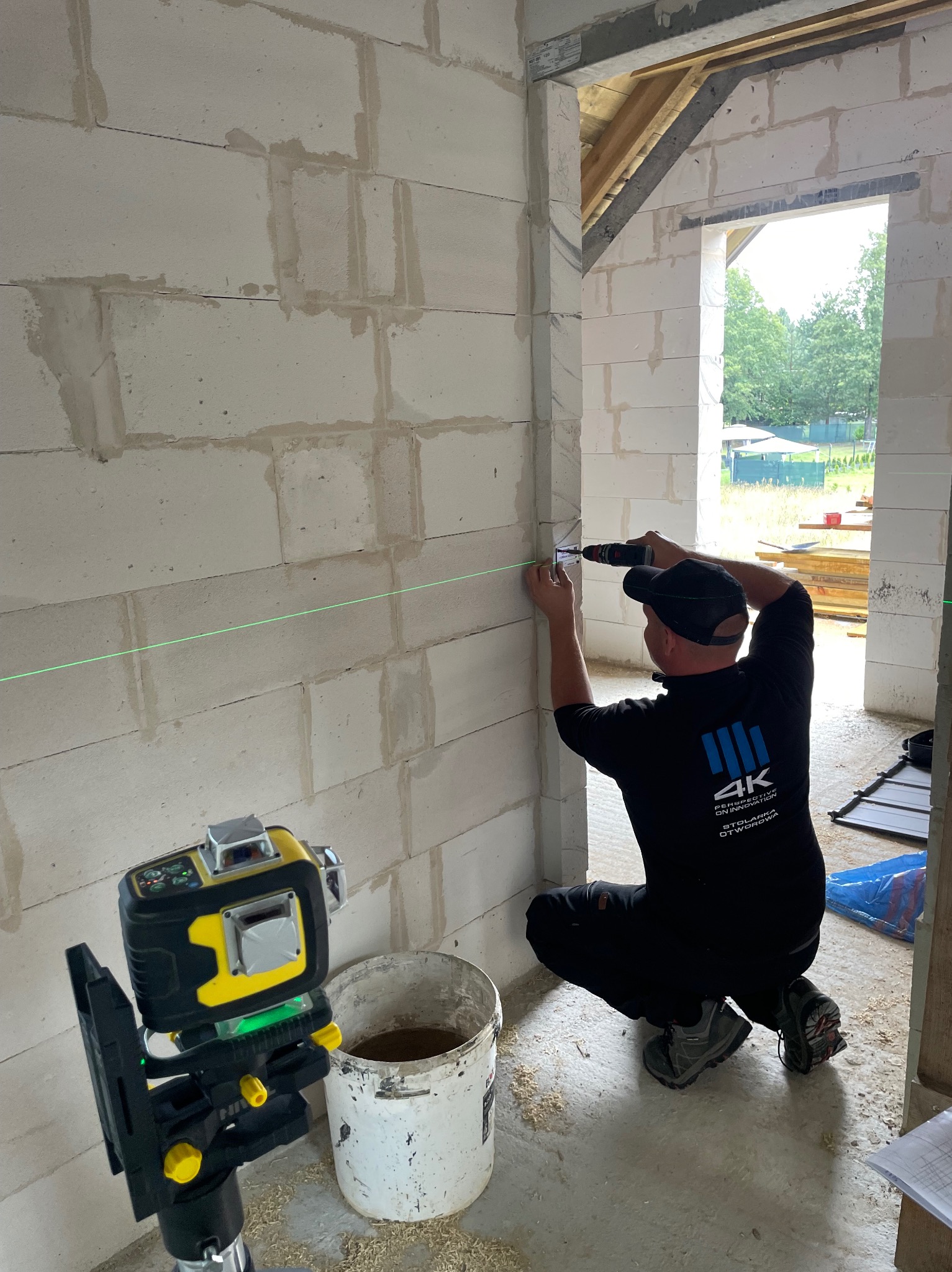 Pracownik w czarnym stroju wierci otwór w ścianie z betonu komórkowego, używając poziomicy laserowej do precyzyjnego wyznaczenia linii. Widoczne narzędzia i surowy stan budowy.